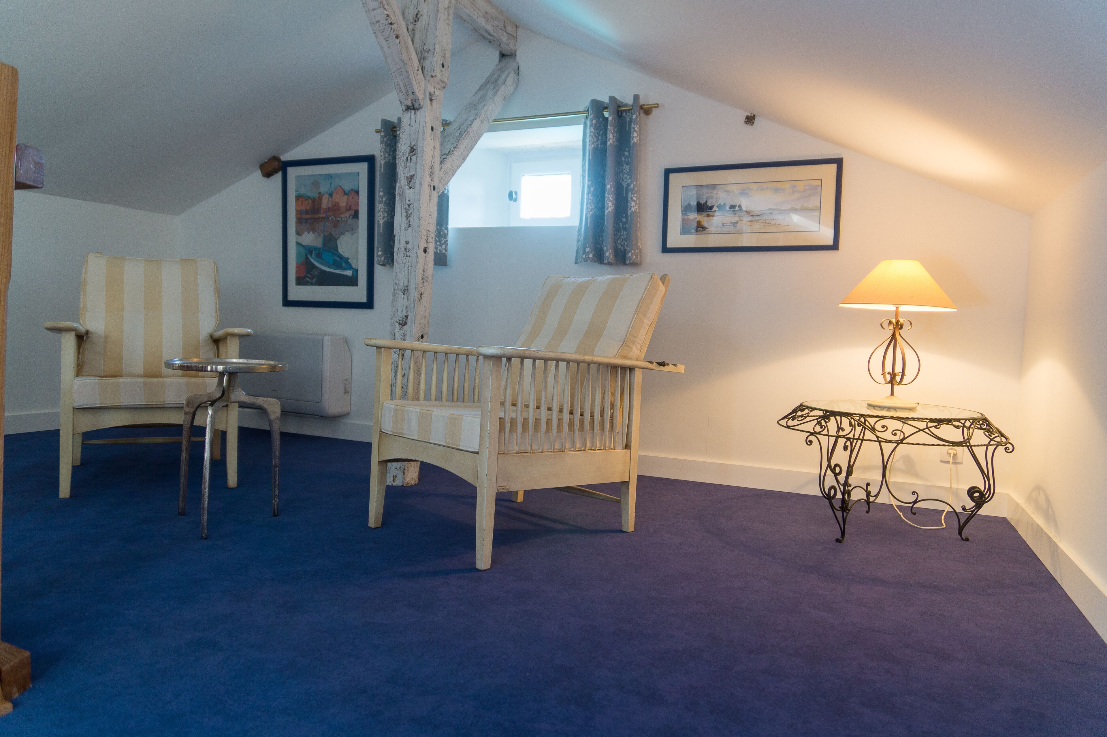 A room with a sloped ceiling, two striped chairs, a small table, a lamp, and two framed pictures on the walls.