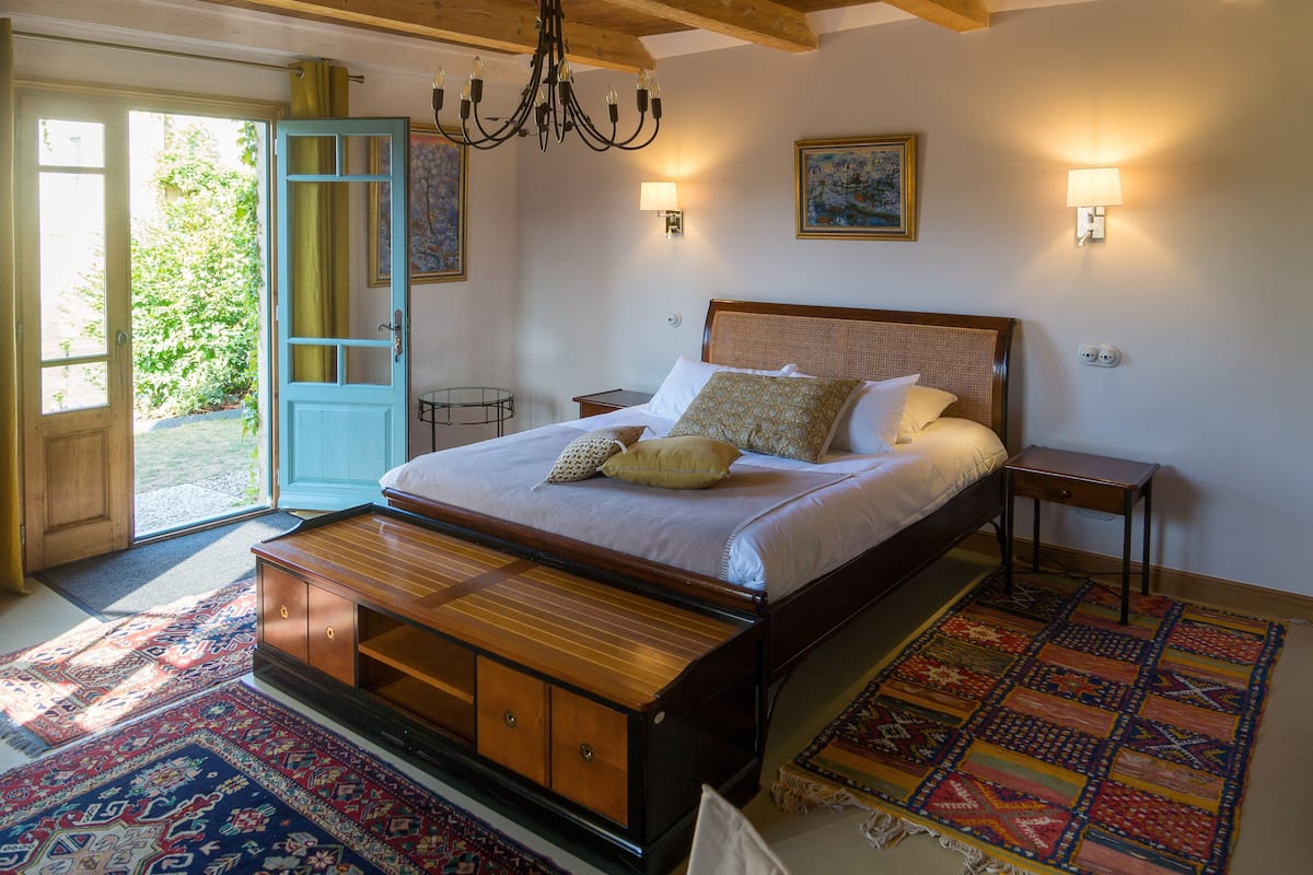 A bedroom with a large bed, wooden headboard, bedside tables, a chandelier, and a colorful rug.