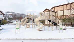 Een speeltuin met glijbanen en klimconstructies, omgeven door een gebouw met een bruine gevel en een met sneeuw bedekte grond.