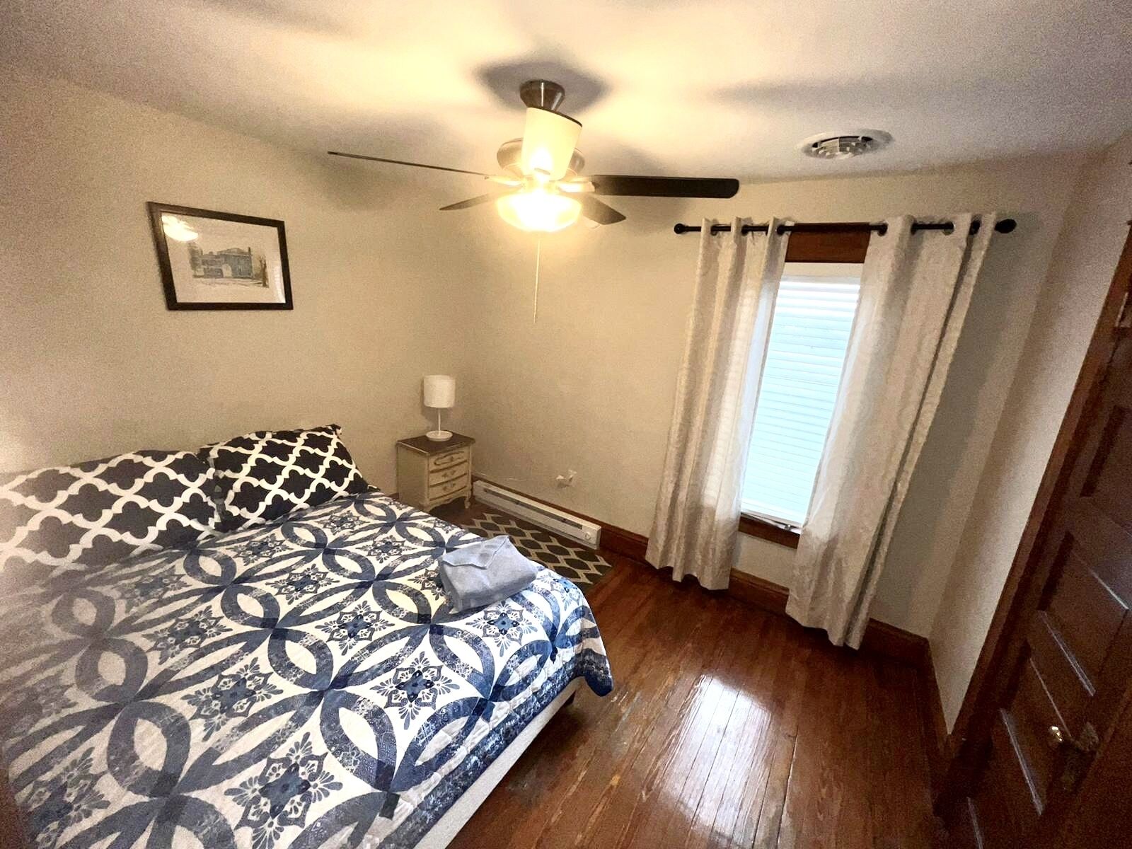 Una camera da letto con un letto, una ventola a soffitto, un comodino, una finestra con tende e un quadro appeso al muro.