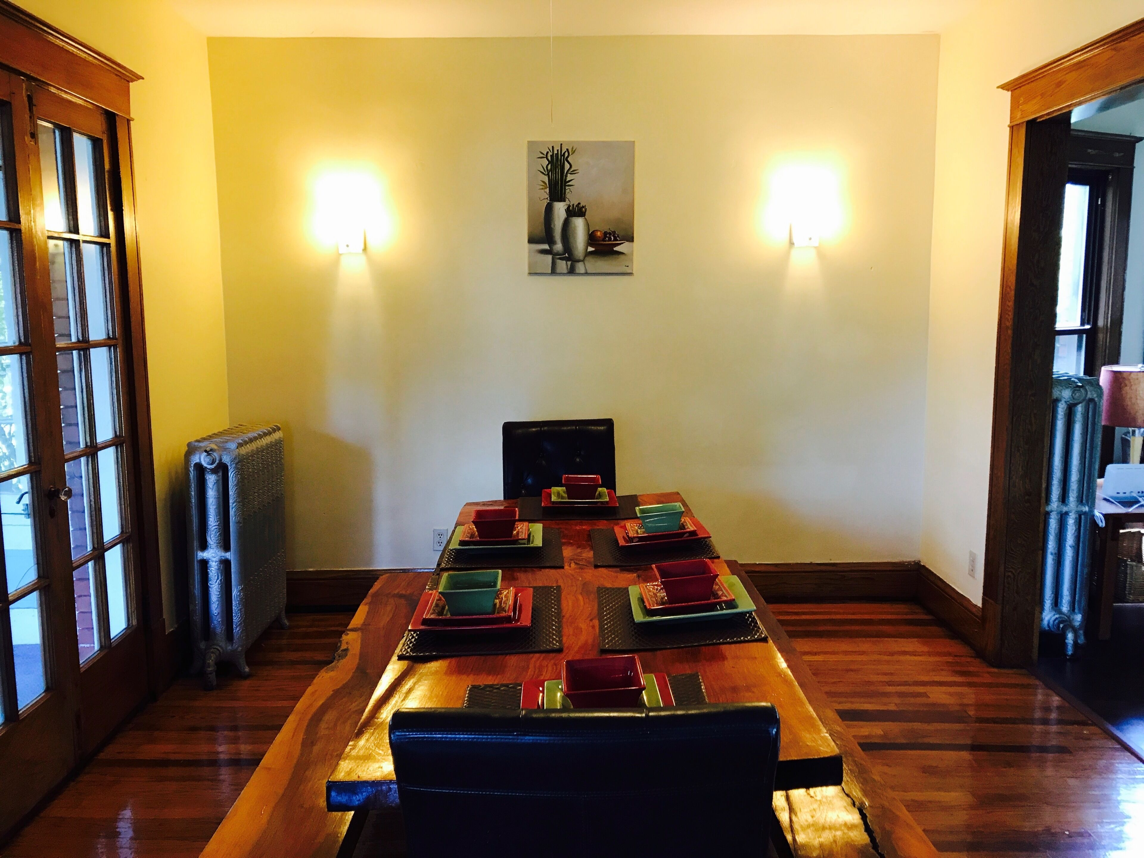 Comedor con una larga mesa de madera dispuesta para una comida, con dos luces fijadas en las paredes y un cuadro encima.