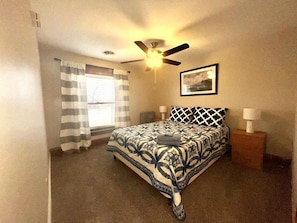 Un dormitorio con cama, ventilador de techo, ventana con cortinas, mesita de noche con lámpara y un cuadro en la pared.