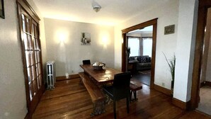Zona pranzo con tavolo e sedie in legno, un termosifone e vista su un soggiorno con divano e una pianta.