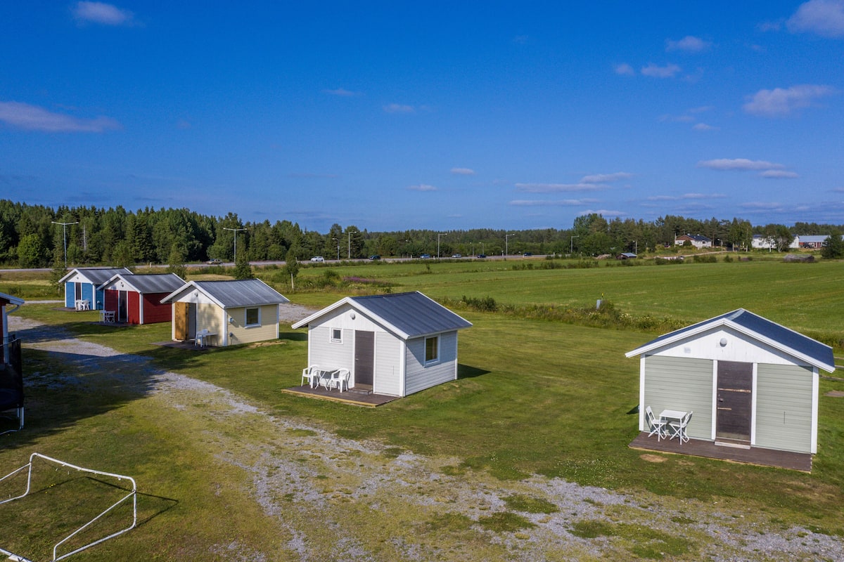 A row of small, single-story cabins with a covered patio area, set in a grassy field with a clear sky.