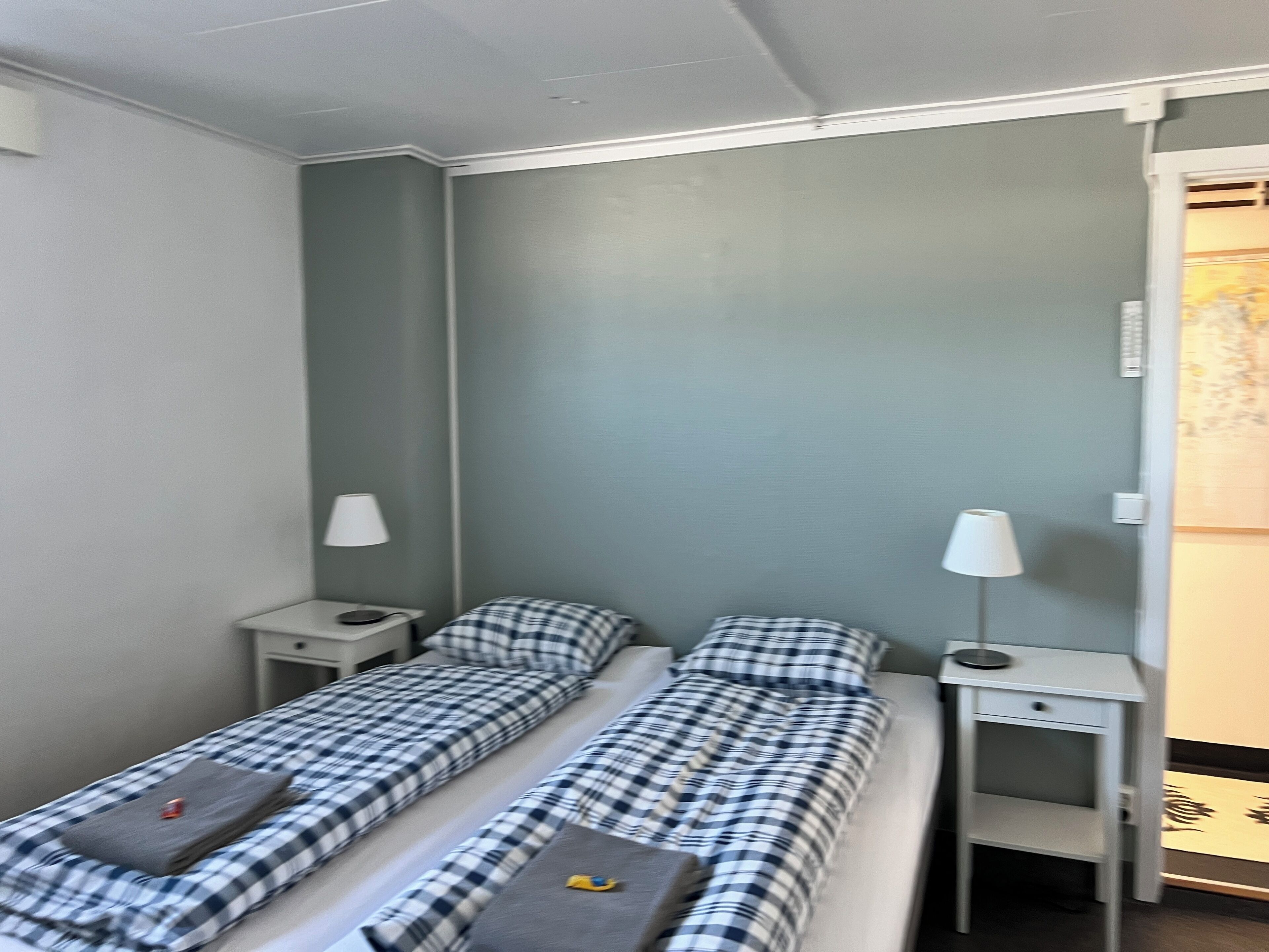 A room with two beds, each with a checkered blanket and pillows, flanked by nightstands with lamps.