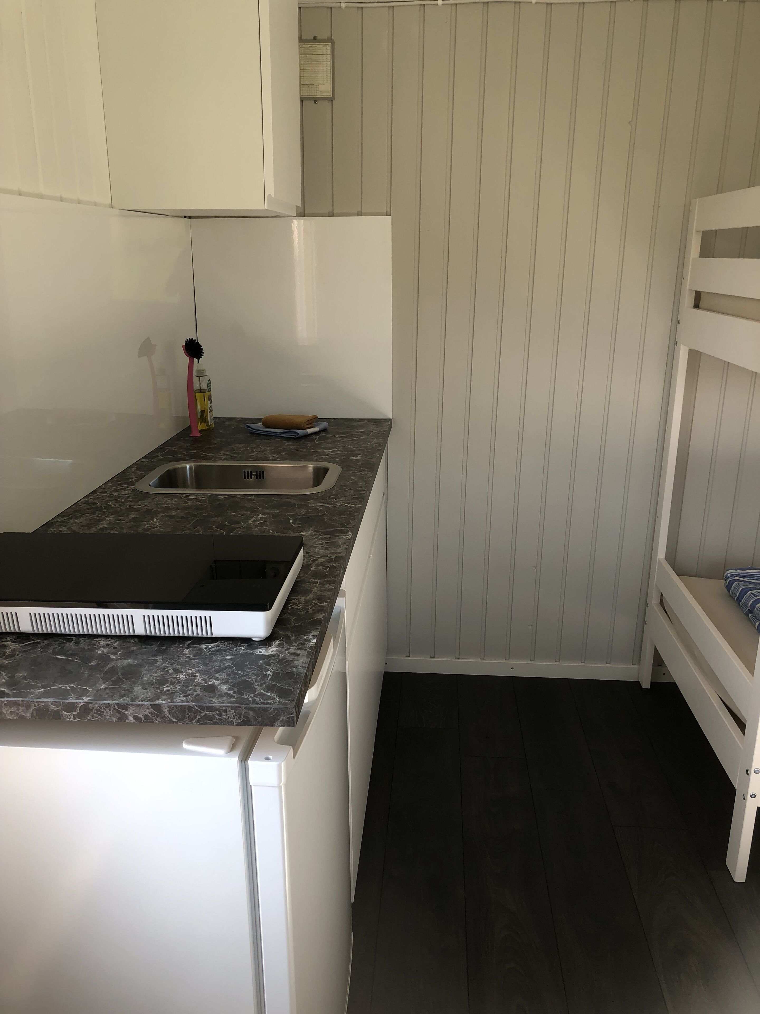 A compact kitchen with a sink, a countertop with a notebook and a bottle, a small white cabinet, and a bunk bed in the corner.