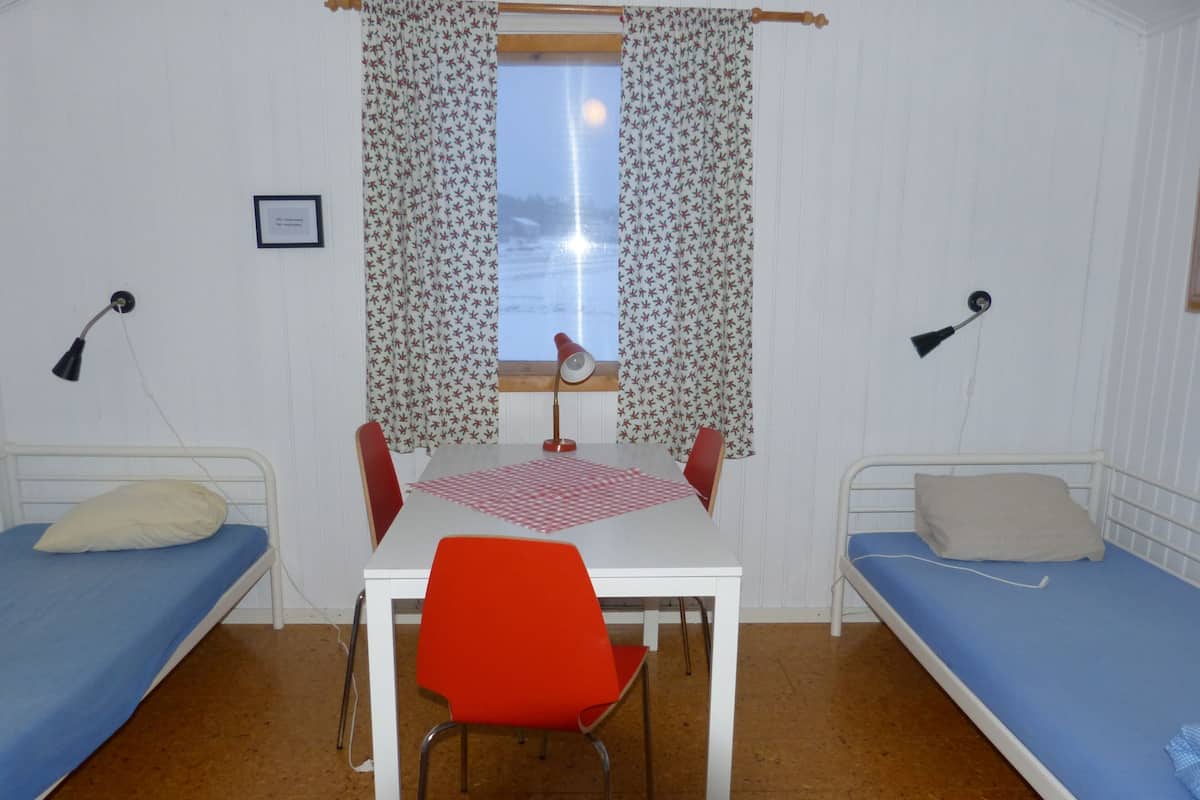 A small, single-room accommodation with two single beds, a table with a chair, a window with curtains, and two bedside lamps.