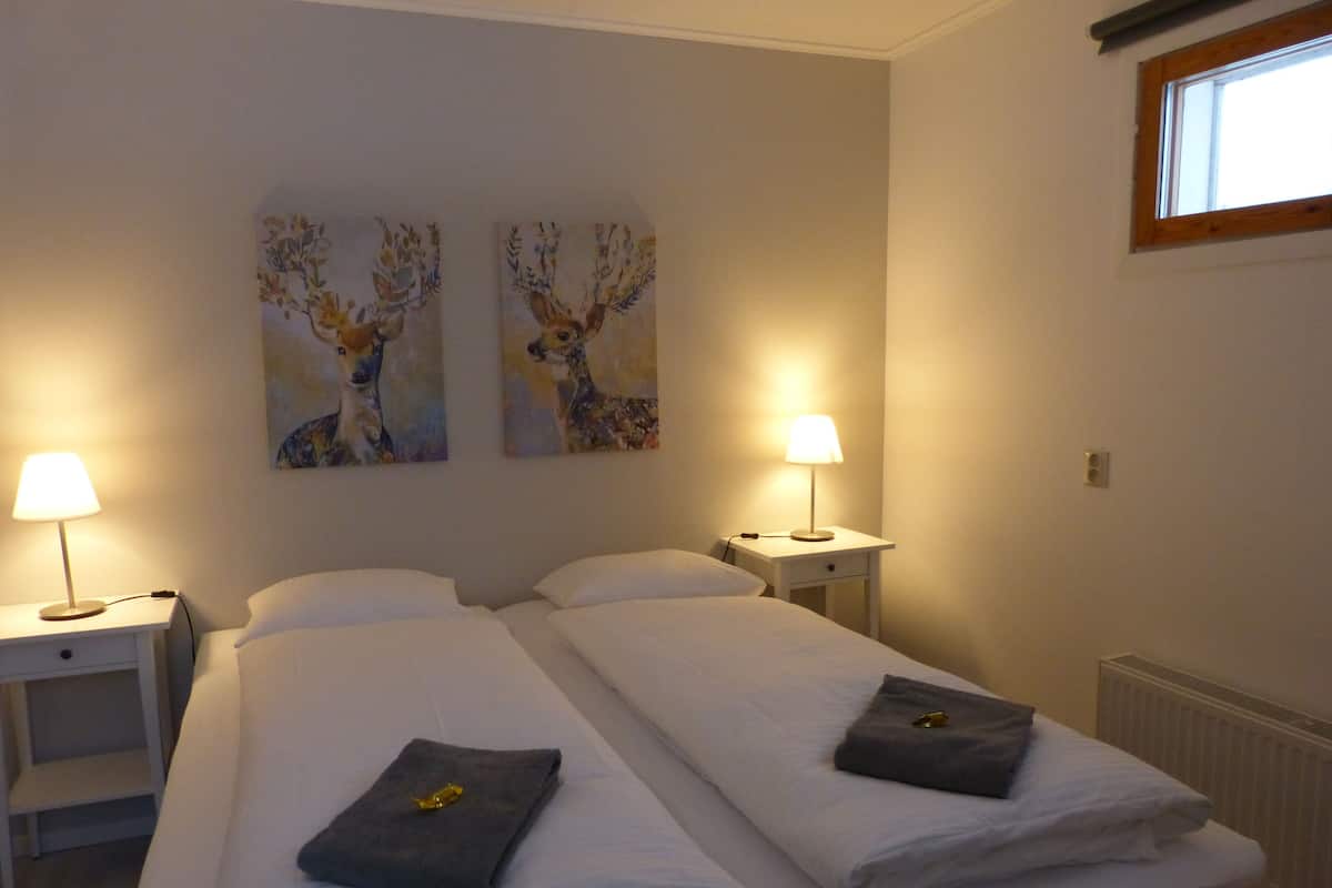 A hotel room with two beds, each with a white duvet and a grey towel. There are two bedside tables with lamps, and two wall-mounted paintings of deer.