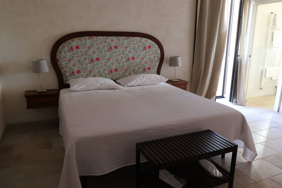 Un lit bien fait, agrémenté d’une tête de lit à motifs floraux, de deux lampes de chevet et d’une petite table sur laquelle se trouve un sac.
