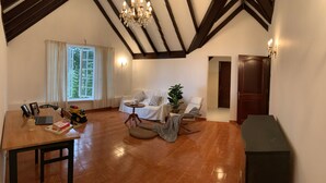 A spacious living area with a wooden table, a sofa, and a chair. There is a window with curtains, a chandelier, and a door. - Green Acres Fraser's Hill (Raub)