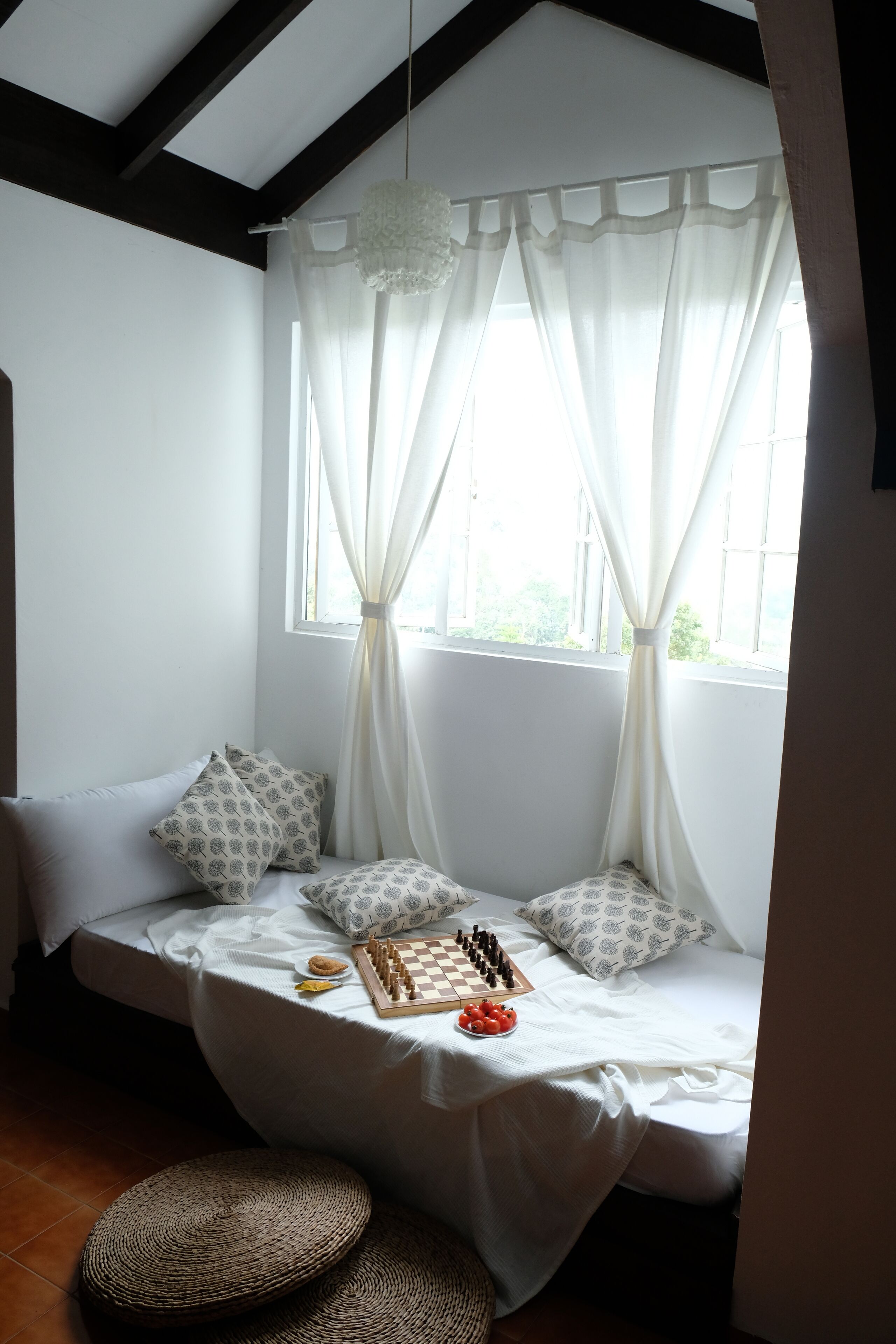 Una habitación acogedora con una cama, una ventana con cortinas traslúcidas, un tablero de ajedrez y una cesta tejida.