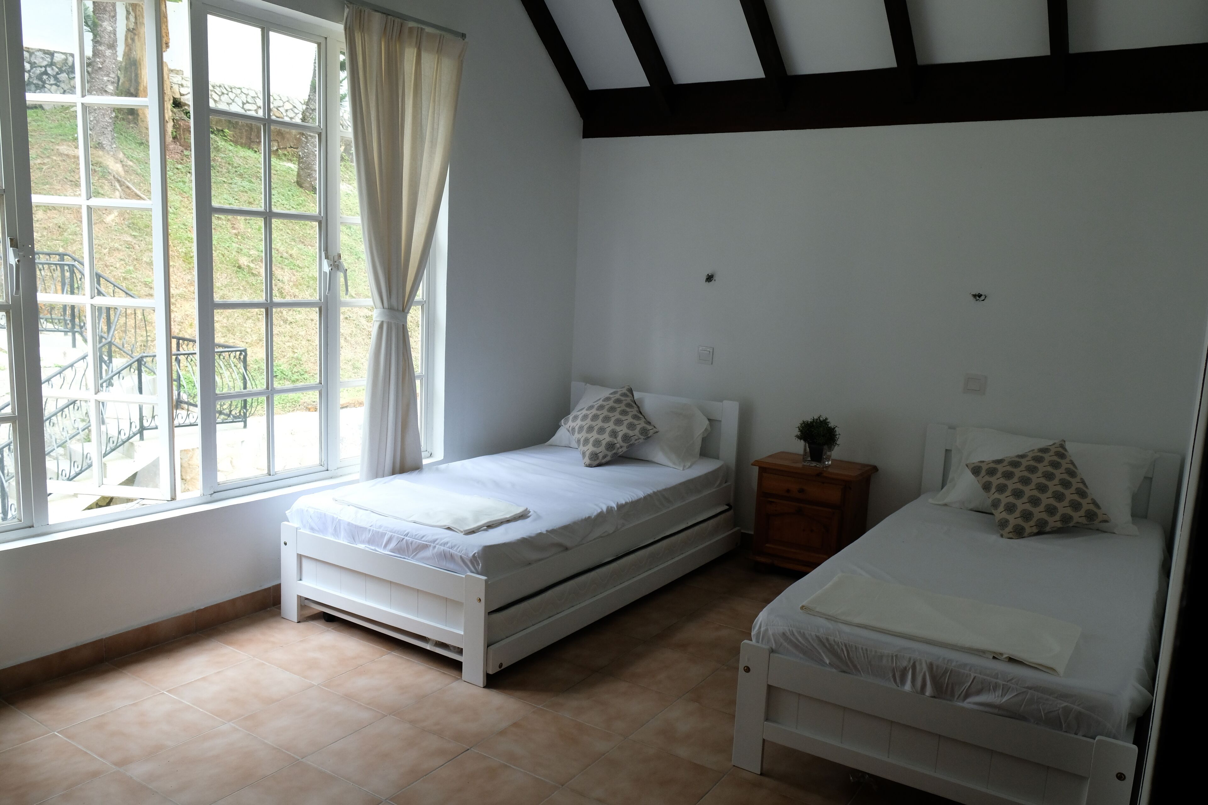 A bedroom with two single beds, a window with curtains, a wooden nightstand, and a view of the outdoors.