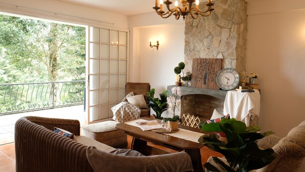 A cozy living room with a fireplace, wicker furniture, and a balcony with a view of trees. - Green Acres Fraser's Hill (Raub)