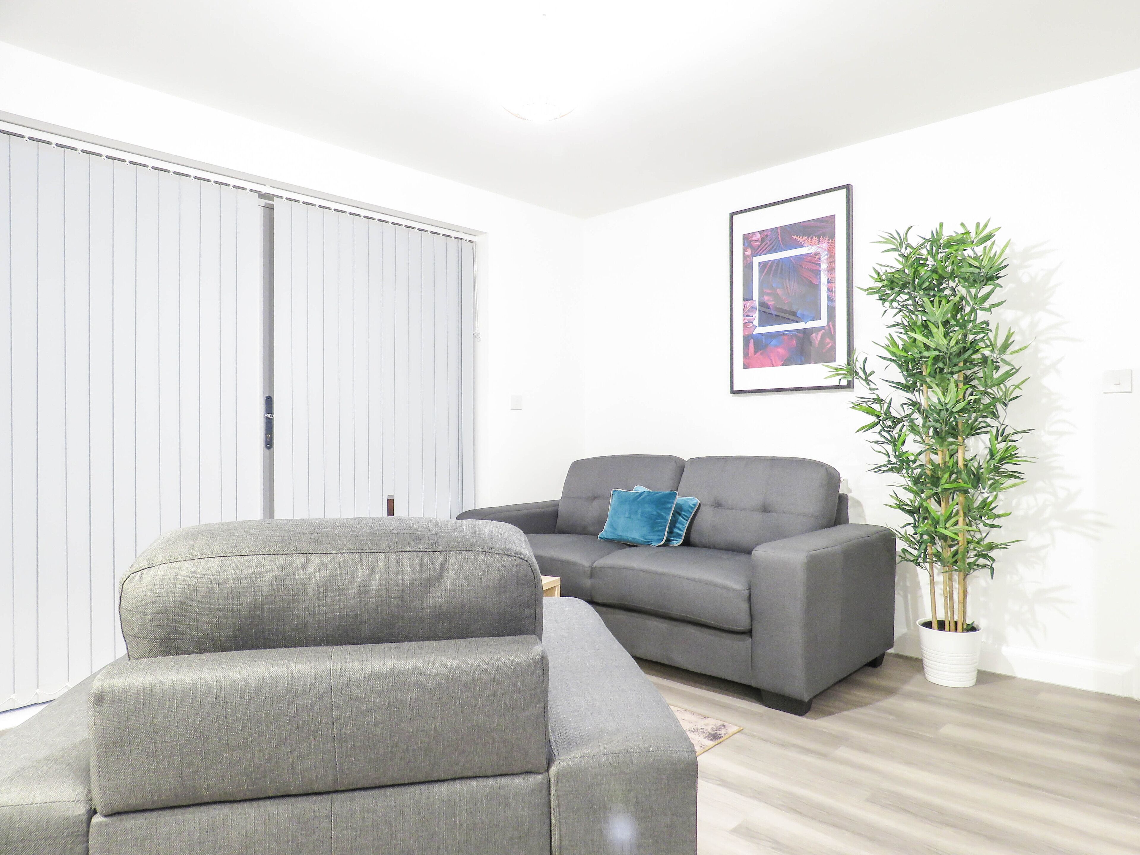 A living room with a grey sofa, a grey armchair, a blue pillow, a framed picture on the wall, and a tall green plant.