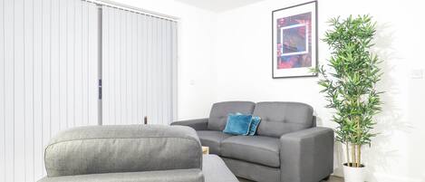 A living room with a grey sofa, a grey armchair, a blue pillow, a framed picture on the wall, and a tall green plant.