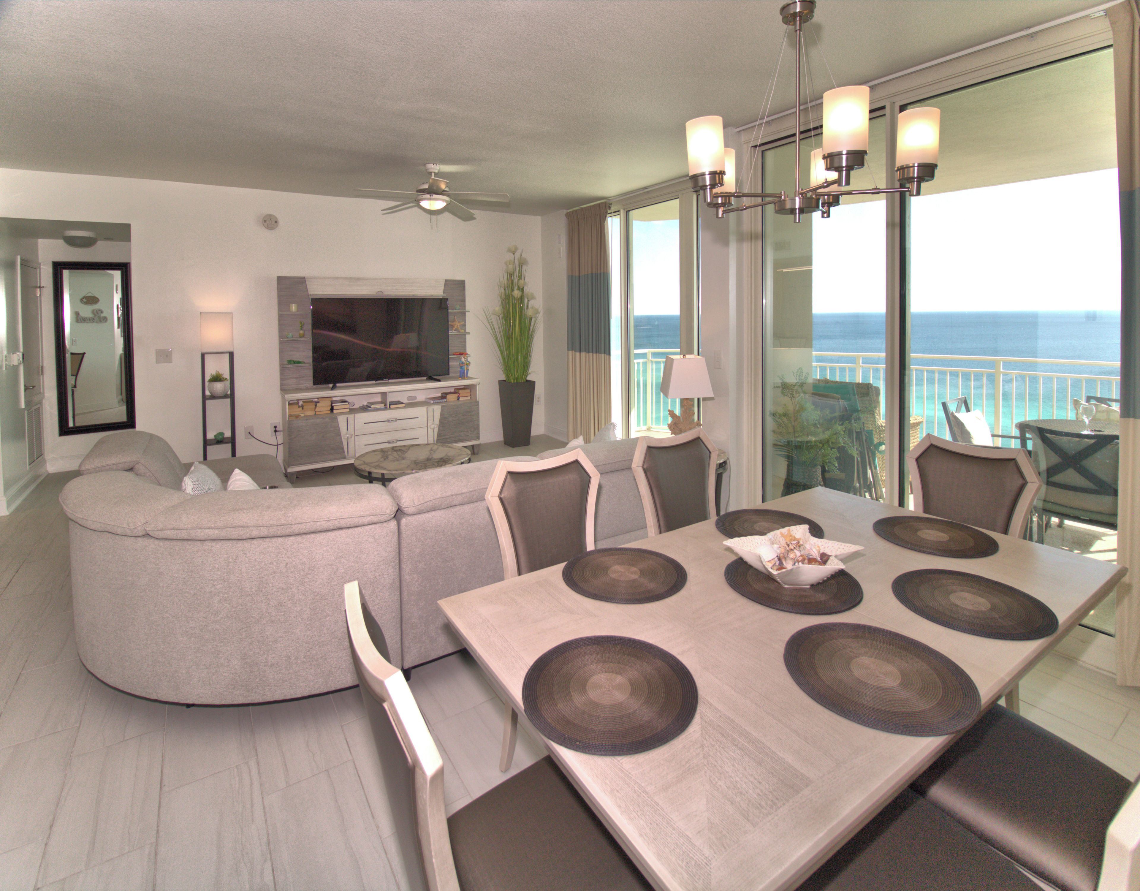 A modern living room with a curved sofa, a dining table with six chairs, a flat-screen TV, and a large window overlooking the ocean.