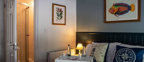 Quarto de hotel com uma cama, uma mesa de cabeceira com abajur, uma ilustração emoldurada de peixe e uma porta para um banheiro.