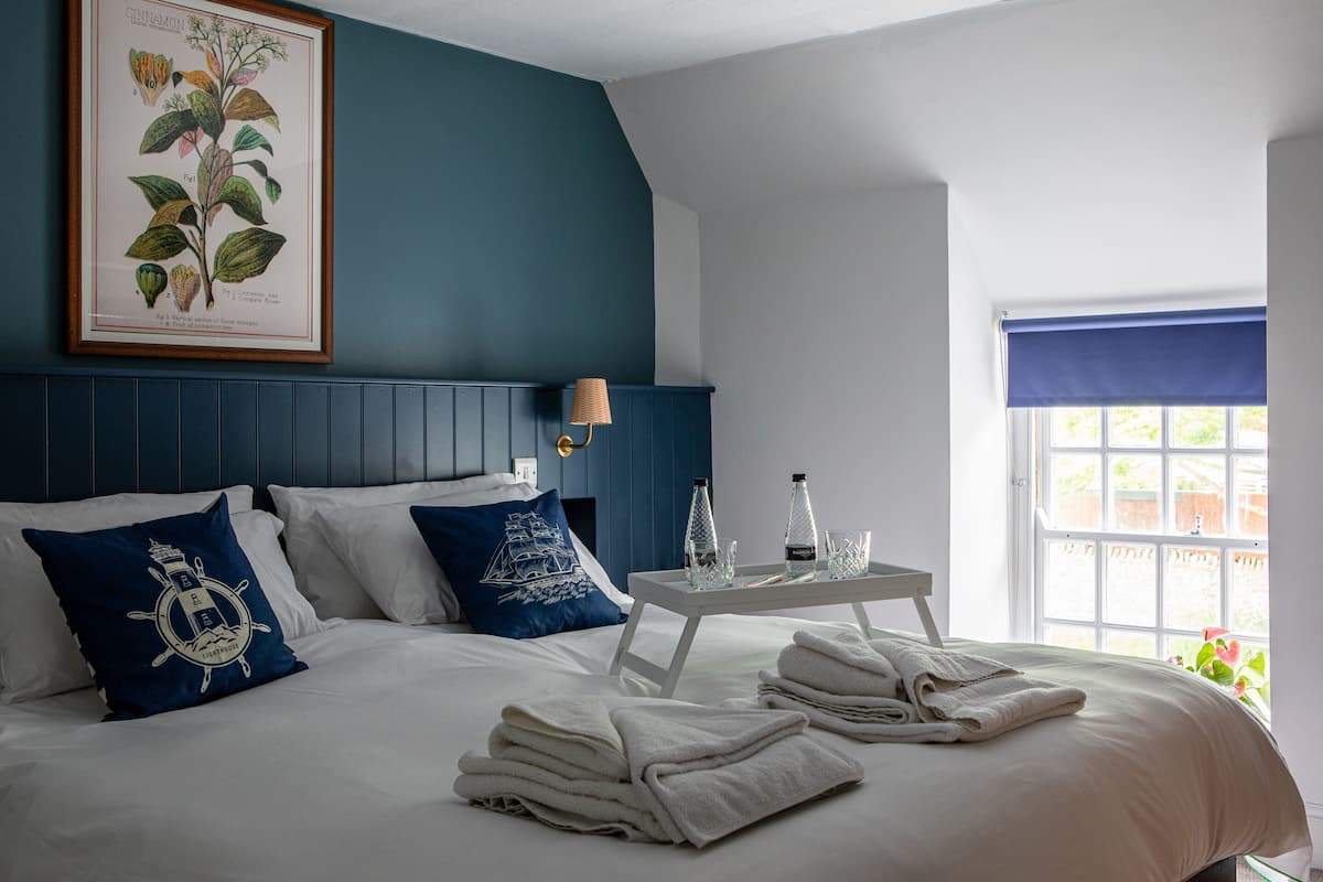 A neatly made bed with white linens and blue pillows, a framed botanical print on the wall, a small table with a lamp and glasses, and a window with a blue curtain.