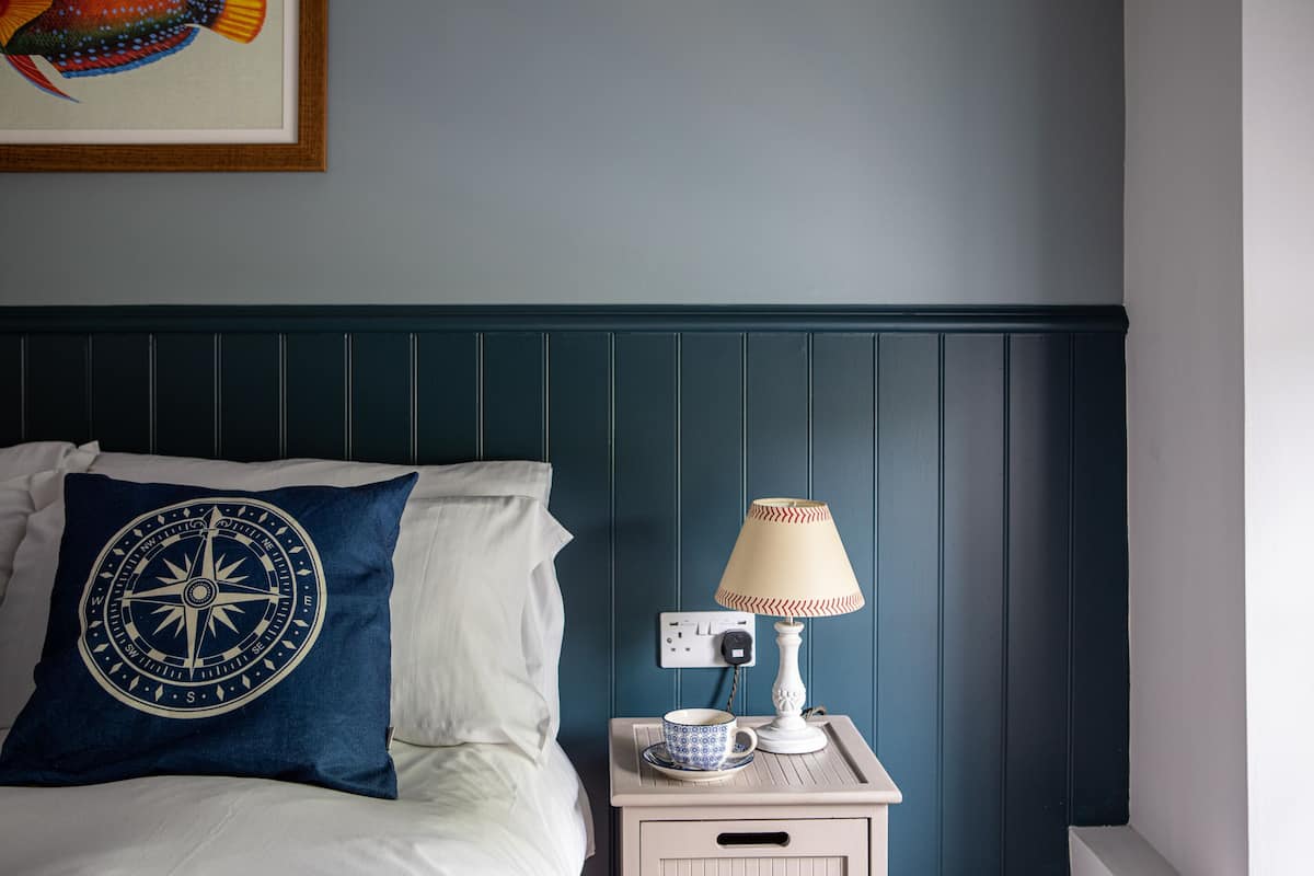 A neatly made bed with a compass-themed pillow, a bedside table with a lamp and a cup, and a wall-mounted power outlet.