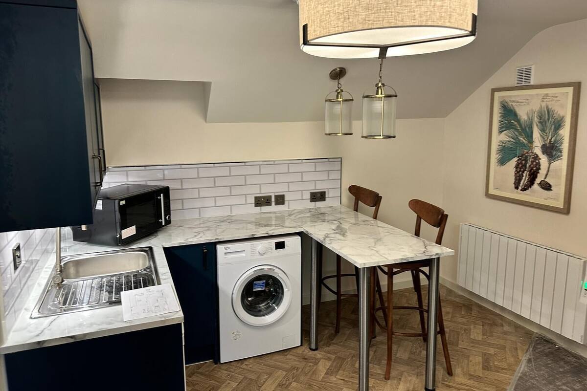 A compact kitchen with a washing machine, a microwave, and a sink. There are two bar stools and a pendant light fixture above the counter.