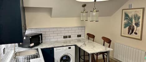 A compact kitchen with a washing machine, a microwave, and a sink. There are two bar stools and a pendant light fixture above the counter.