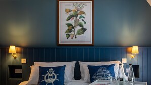 A hotel room with a bed, two pillows with nautical designs, a framed botanical illustration, bedside lamps, and a tray with glasses and a bottle. - The Spice Ship (Weymouth)