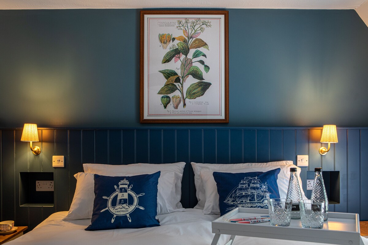 A hotel room with a bed, two pillows with nautical designs, a framed botanical illustration, bedside lamps, and a tray with glasses and a bottle.