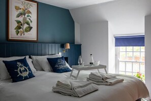 A neatly made bed with white linens and blue pillows, a framed botanical print on the wall, a small table with glasses and bottles, and a window with a blue blind. - The Spice Ship (Weymouth)