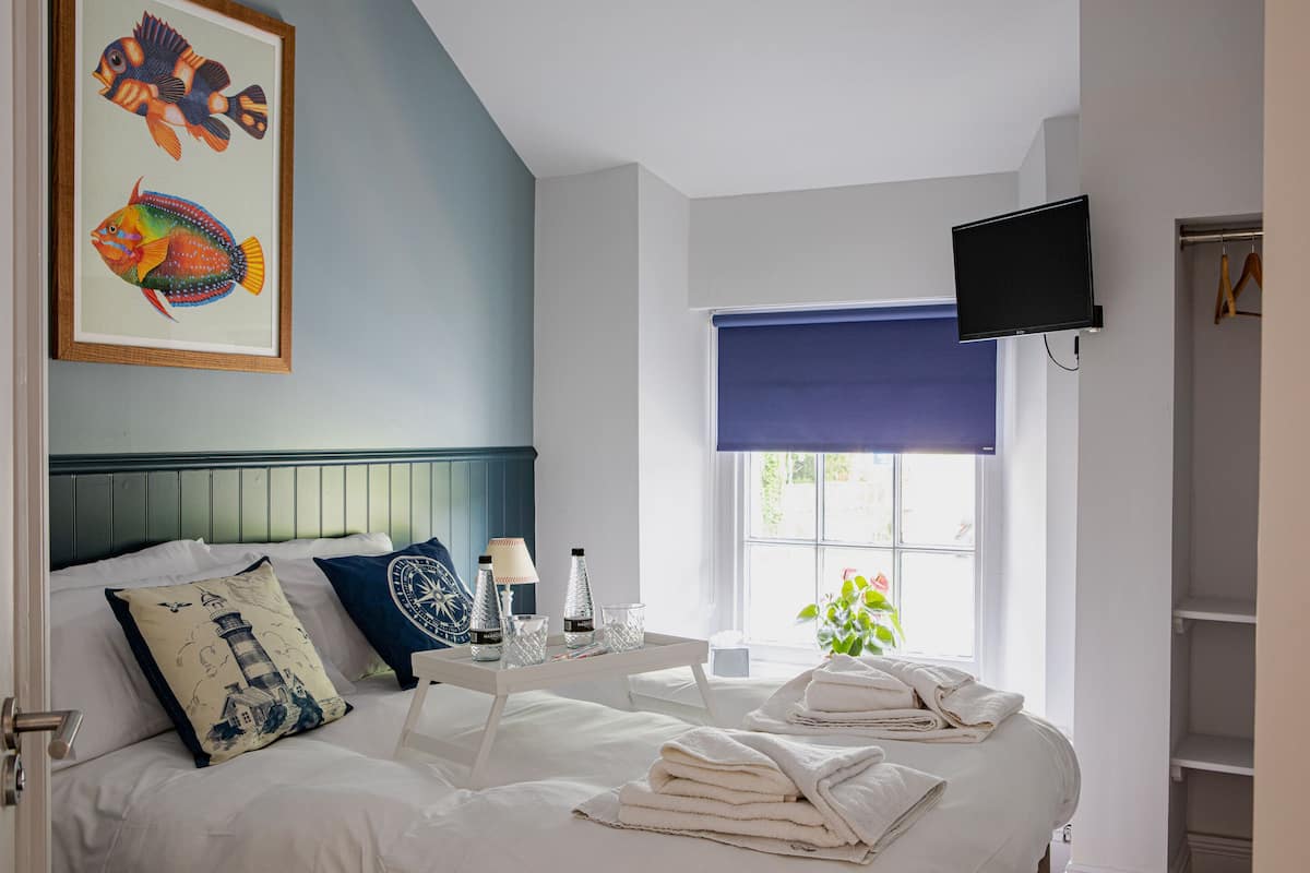 A hotel room with a bed, a TV mounted on the wall, a window with a blue blind, and a painting of fish on the wall.