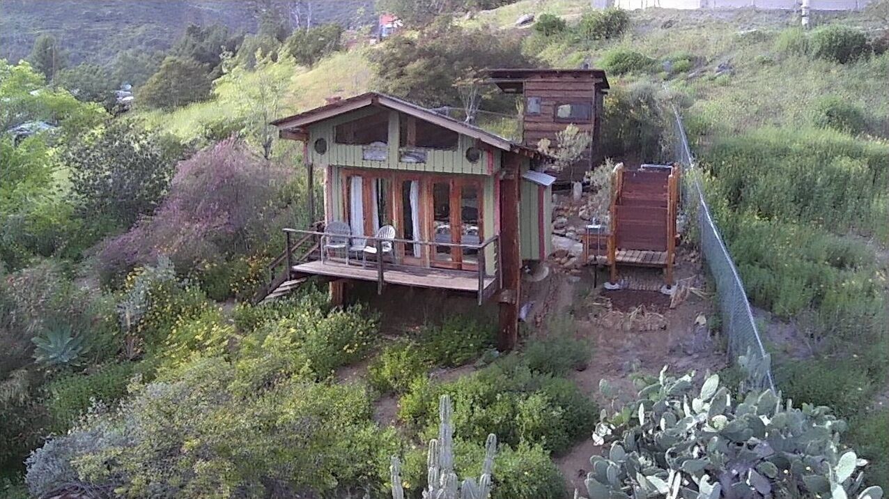 A rustic cabin with a balcony, surrounded by lush vegetation and a steep hillside.