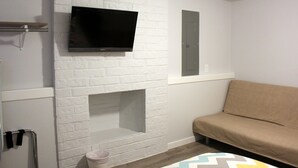 A room with a white brick wall, a mounted flat-screen TV, a sofa, and a small wooden table. - Rainier Guesthouse (Walk to Metro) (Seattle)