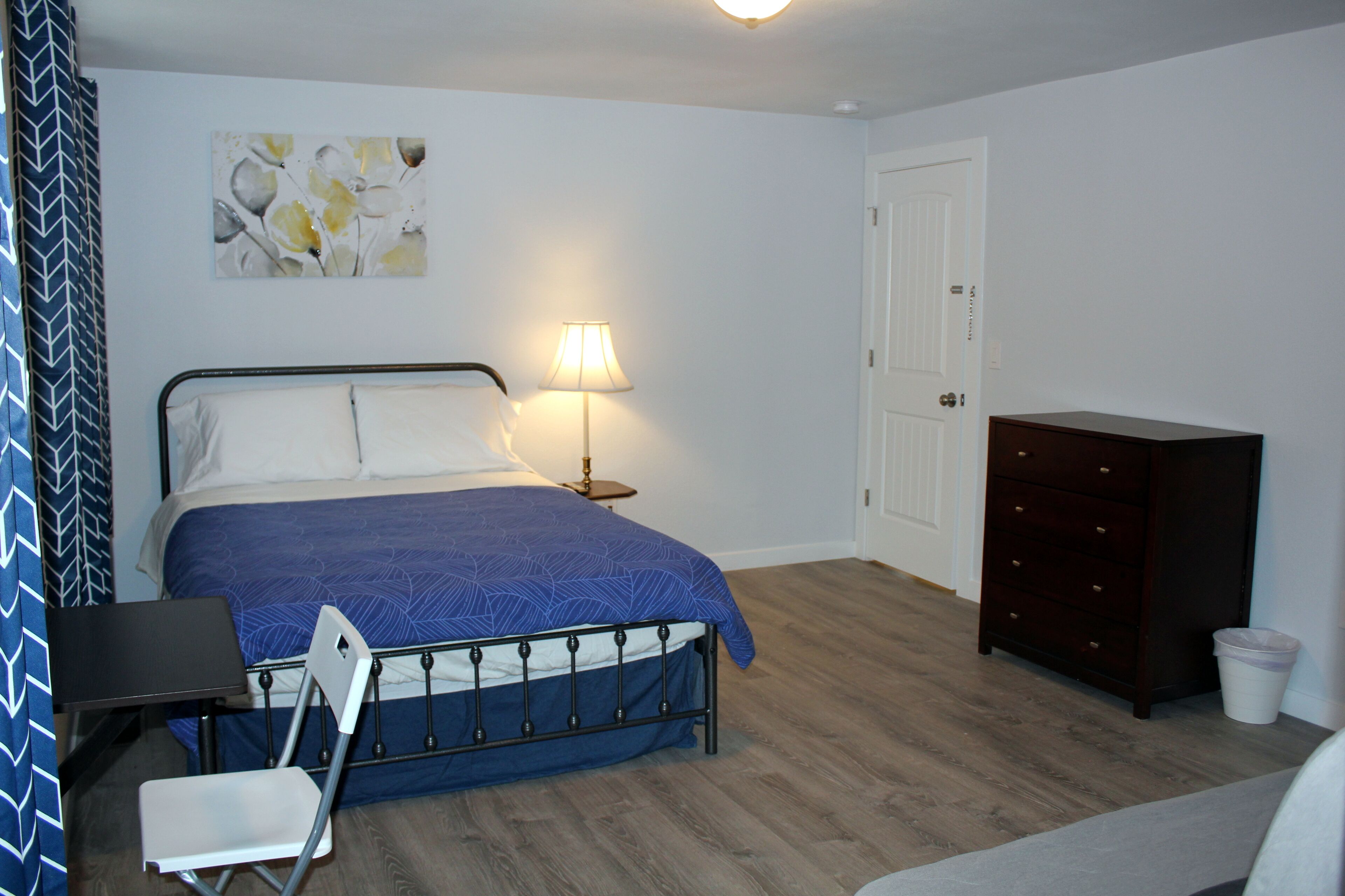A bedroom with a bed, a desk, a chair, a dresser, and a lamp.