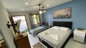 Ein Hotelzimmer mit einem großen Bett, einem kleineren Bett, einem Schreibtisch mit Computer, einem Deckenventilator und einem Gemälde an der Wand.
