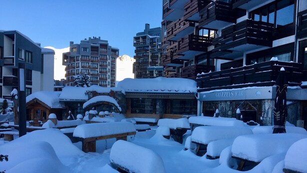 Ein mit Schnee bedeckter Außenbereich mit Bänken und Tischen, ein rustikales Holzgebäude und im Hintergrund eine Bar namens 'Grizzly's Bar'.
