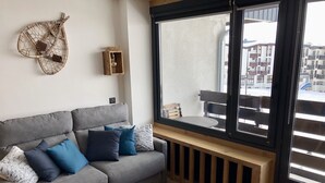 Ein modernes Wohnzimmer mit grauem Sofa, blauen Kissen, einem kleinen Holztisch und einem großen Fenster mit Blick auf einen Balkon.
