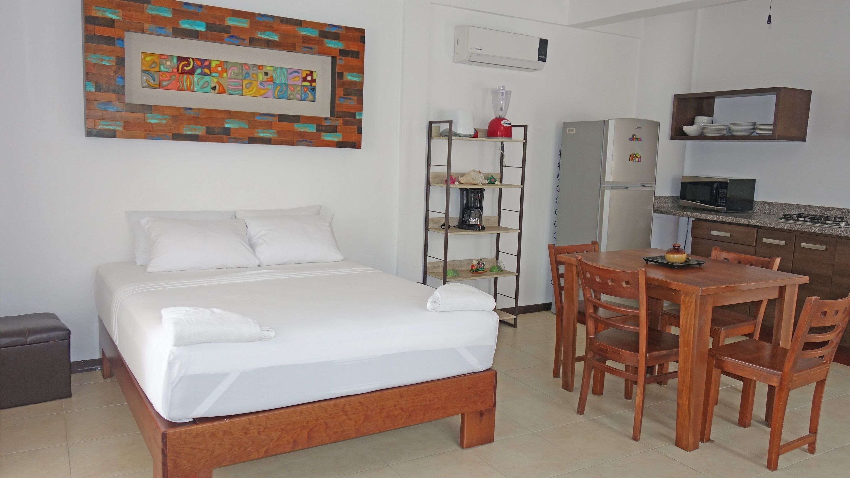 Une chambre d’hôtel comprenant un lit, une table à manger en bois avec des chaises, un coin cuisine avec un réfrigérateur, un four à micro-ondes et une cuisinière, ainsi qu’une étagère contenant des ustensiles de cuisine.