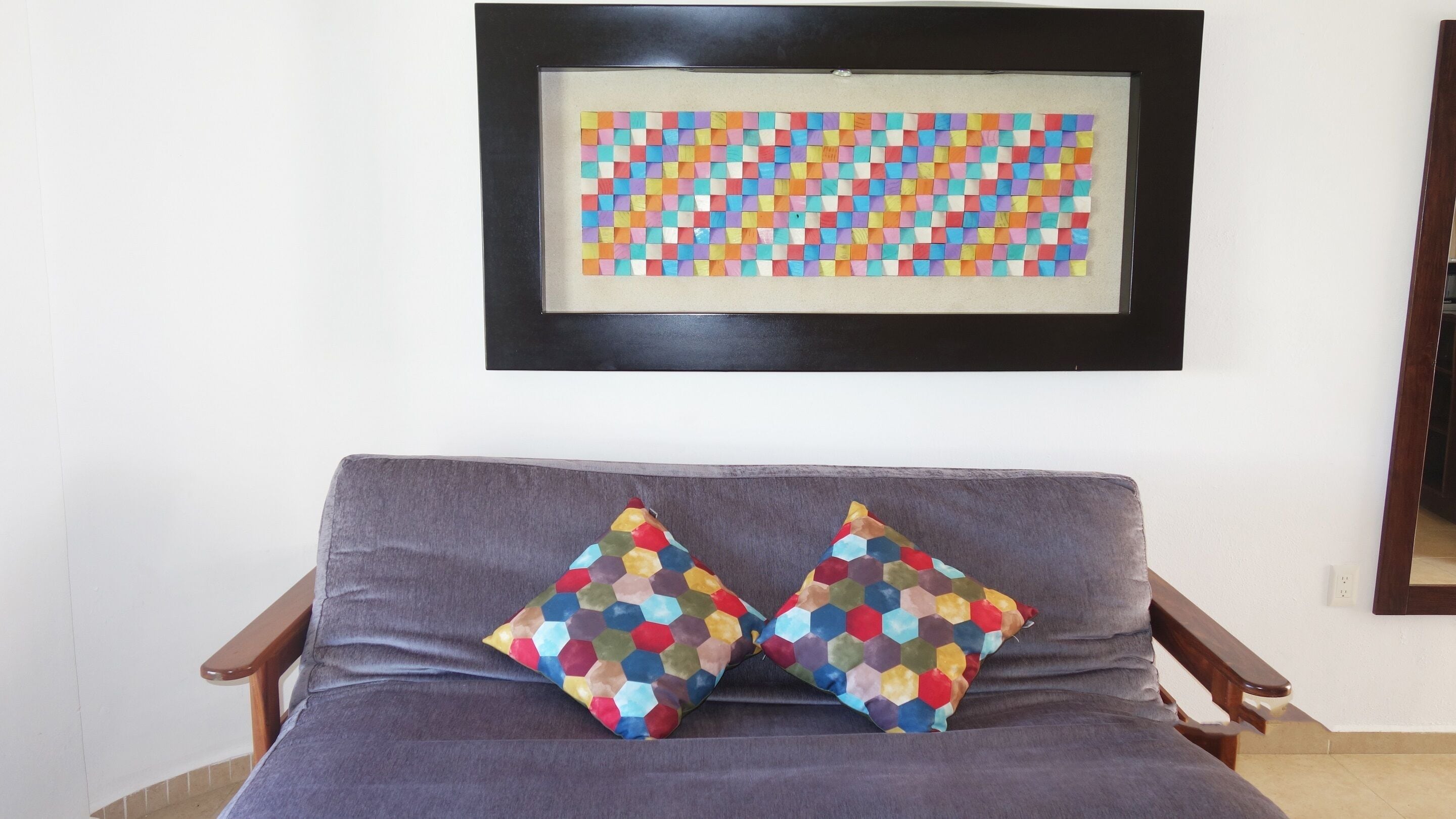 A grey sofa with two colorful hexagonal pillows, a framed artwork above, and a wooden chair to the left.