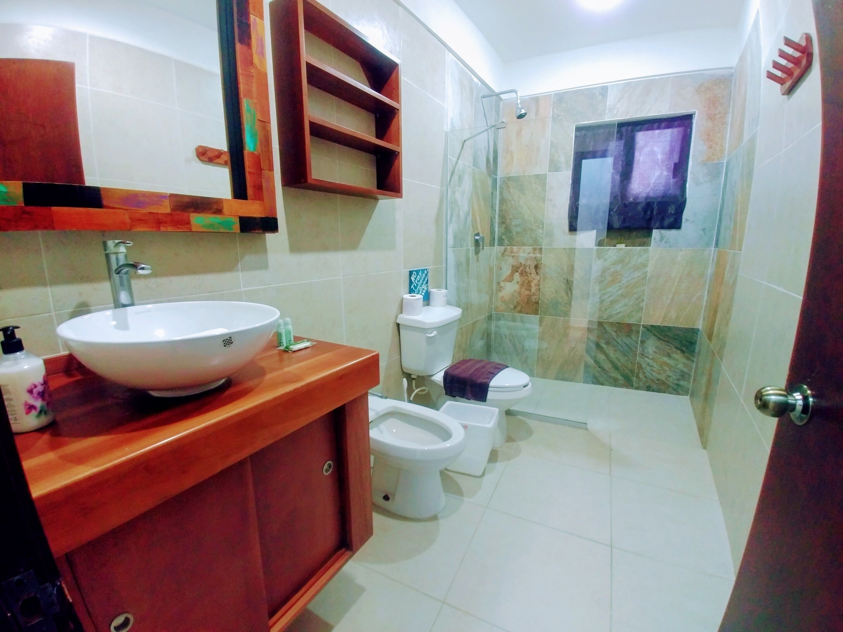 Un bagno con un lavandino grande e bianco, un mobile contenitore in legno, due servizi igienici e una cabina doccia con pareti in vetro.