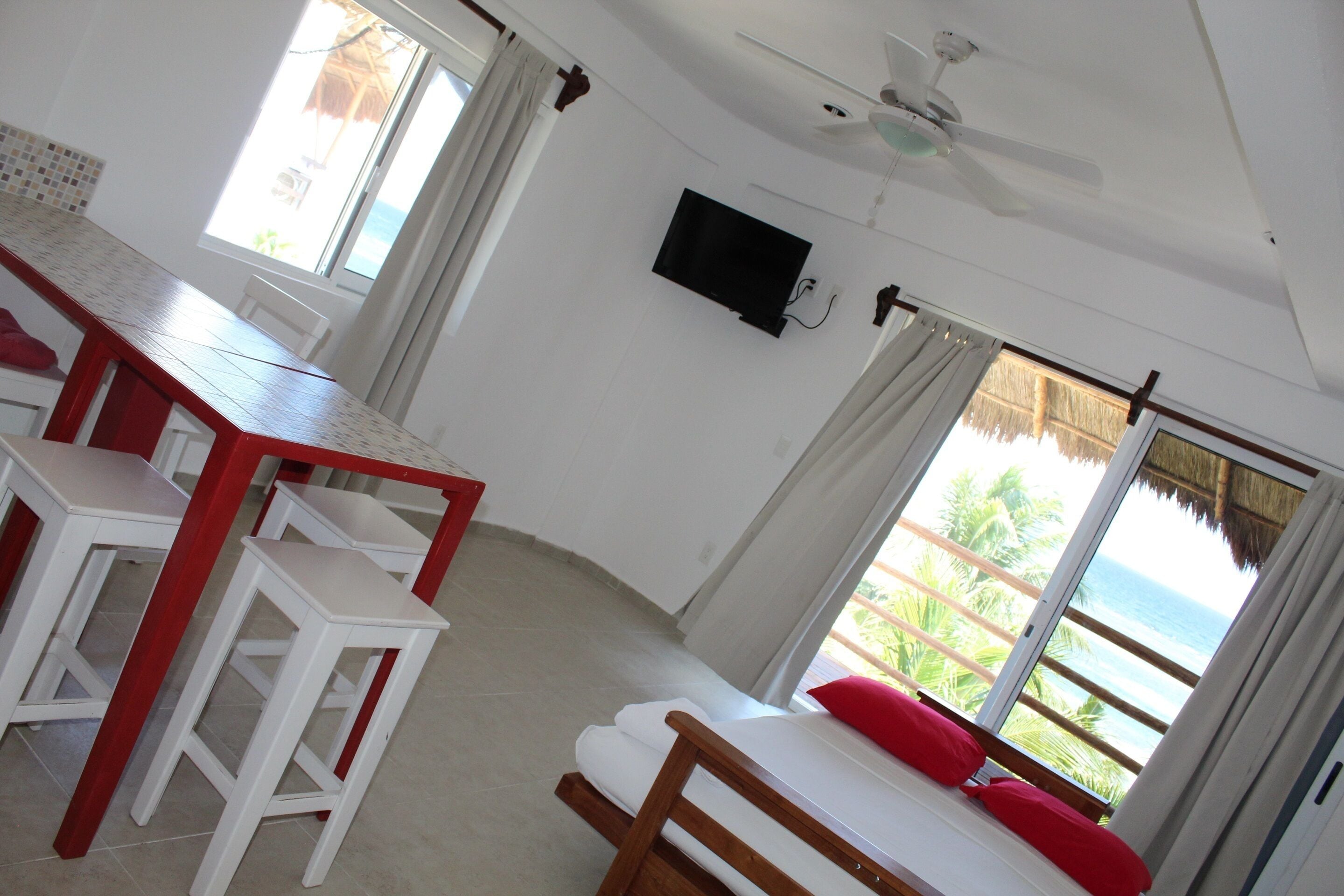 A room with a bed, a TV mounted on the wall, a dining table with chairs, and a view of the ocean through large windows.