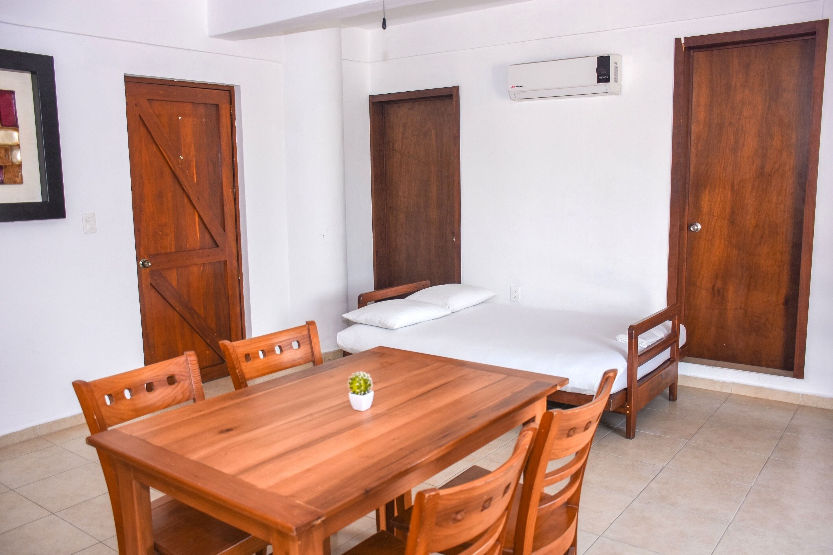 Una habitación con una mesa de comedor de madera, cuatro sillas, una cama con ropa de cama blanca y dos puertas de madera.