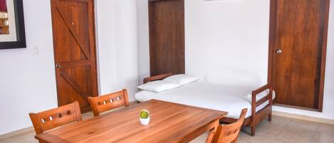 Una habitación con una mesa de comedor de madera, cuatro sillas, una cama con ropa de cama blanca y dos puertas de madera.