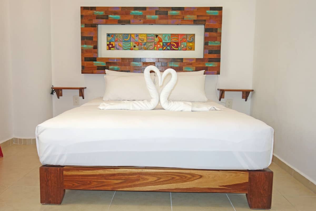 A neatly made bed with white linens and two pillows, a wooden headboard, a colorful abstract painting above the bed, and a red curtain on the left.