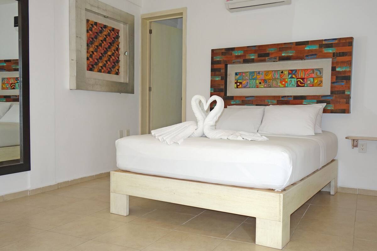 A neatly made bed with a white comforter and pillows, a wooden headboard, a wall-mounted air conditioning unit, a framed abstract art piece above the bed, and a small bedside table with a power outlet.