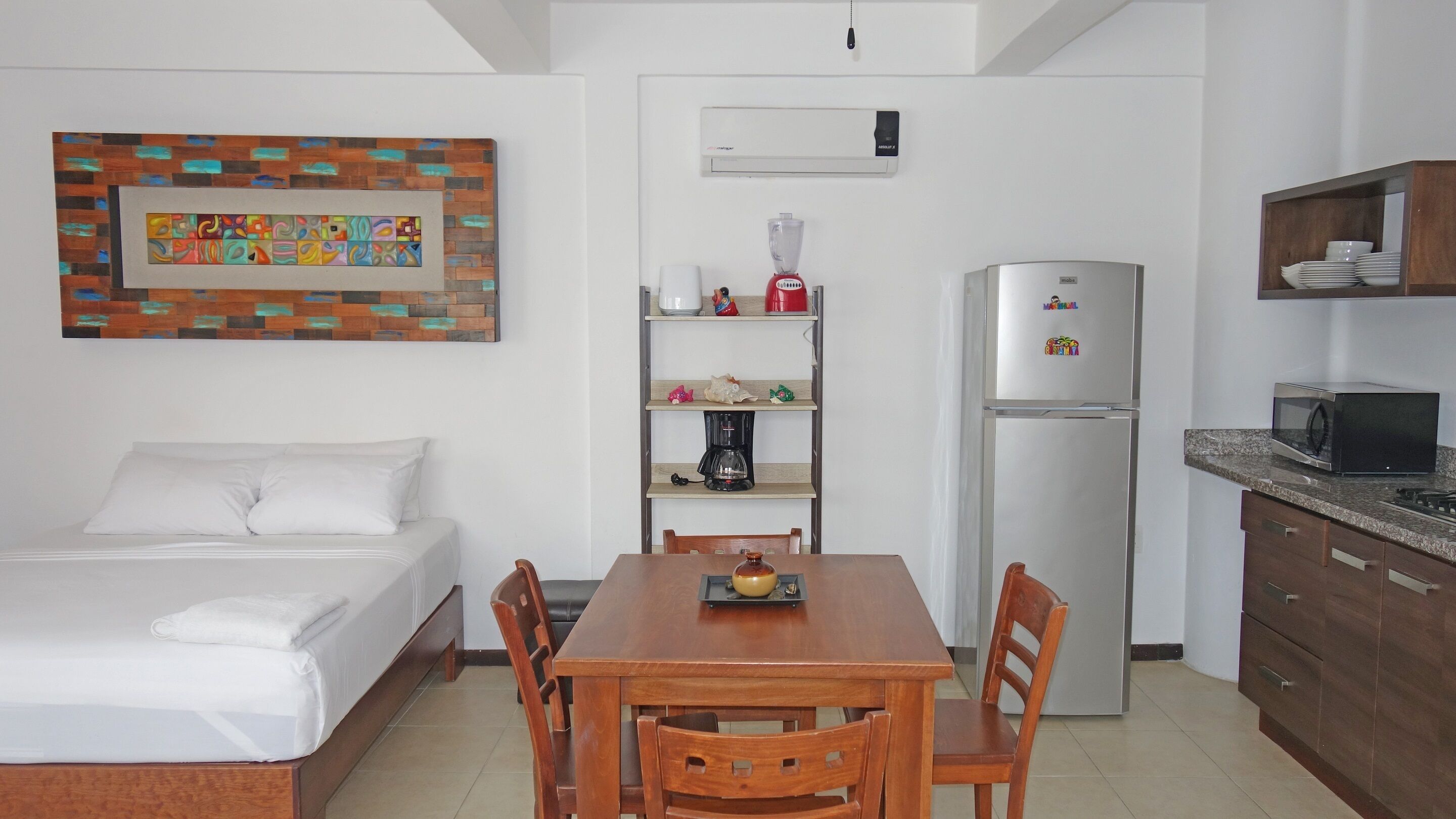 A compact hotel room with a bed, a wooden dining table with chairs, a kitchen area with a refrigerator, microwave, and a coffee maker.