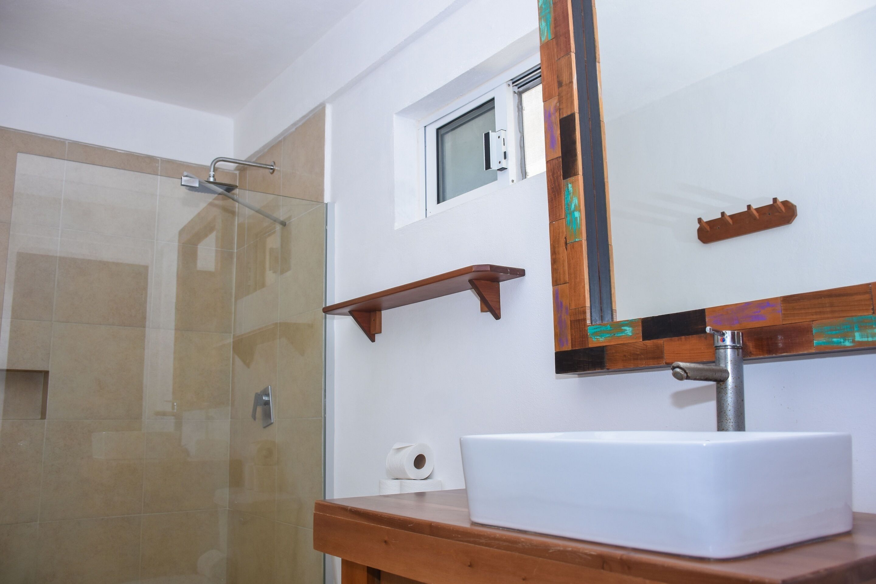 A bathroom with a glass shower enclosure, a white sink, a wooden shelf, and a mirror with a decorative frame.