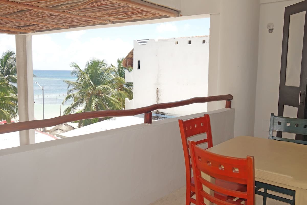 A balcony with a table and chairs, overlooking a beach with palm trees and the ocean.