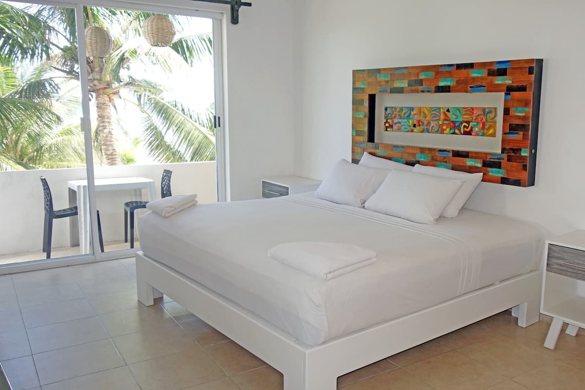 A bedroom with a large bed, a bedside table, a chair, and a view of palm trees through a sliding glass door.