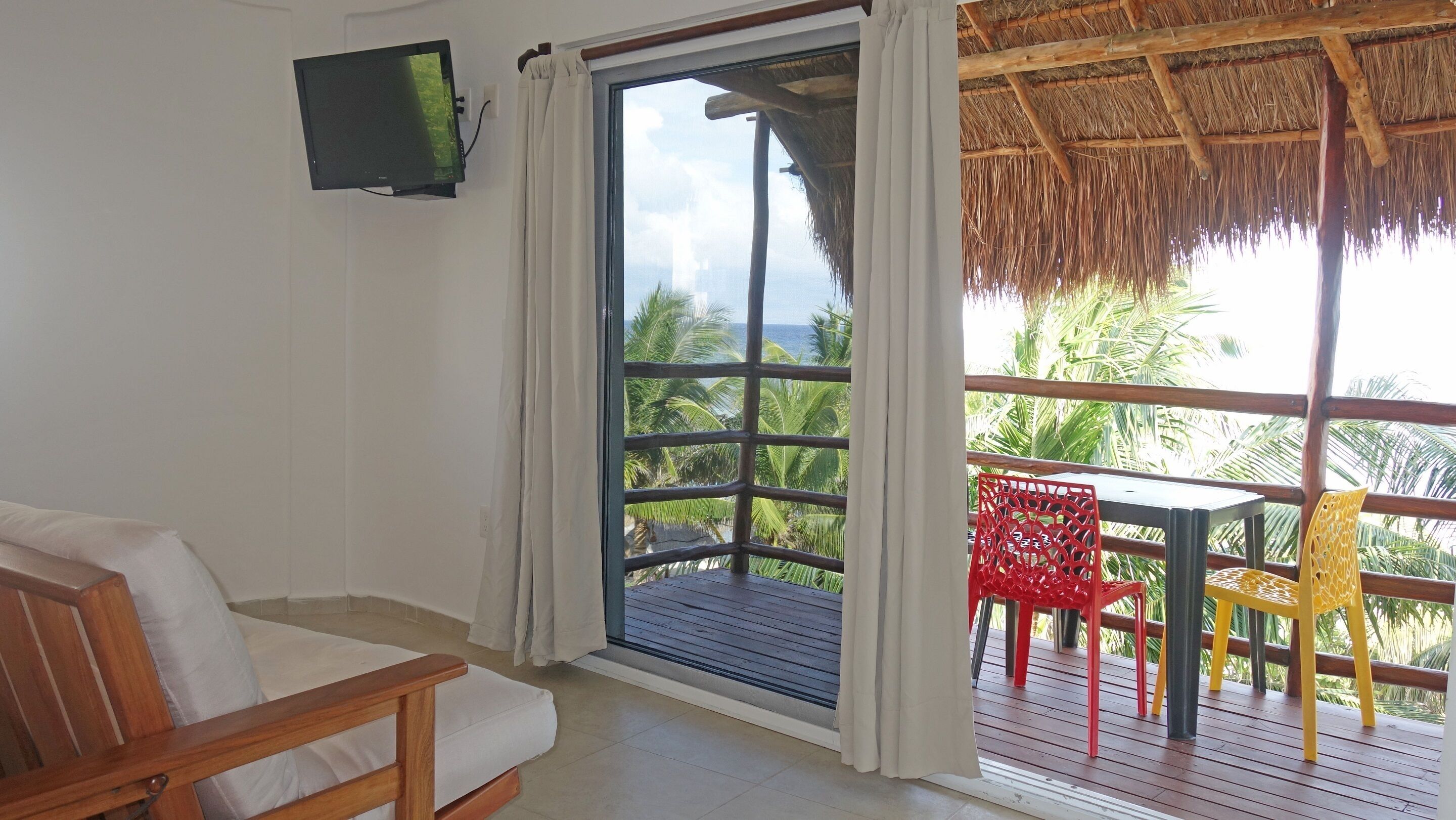 Una habitación con una cama de madera, un televisor montado en la pared y un ventanal con vistas a palmeras y al océano.