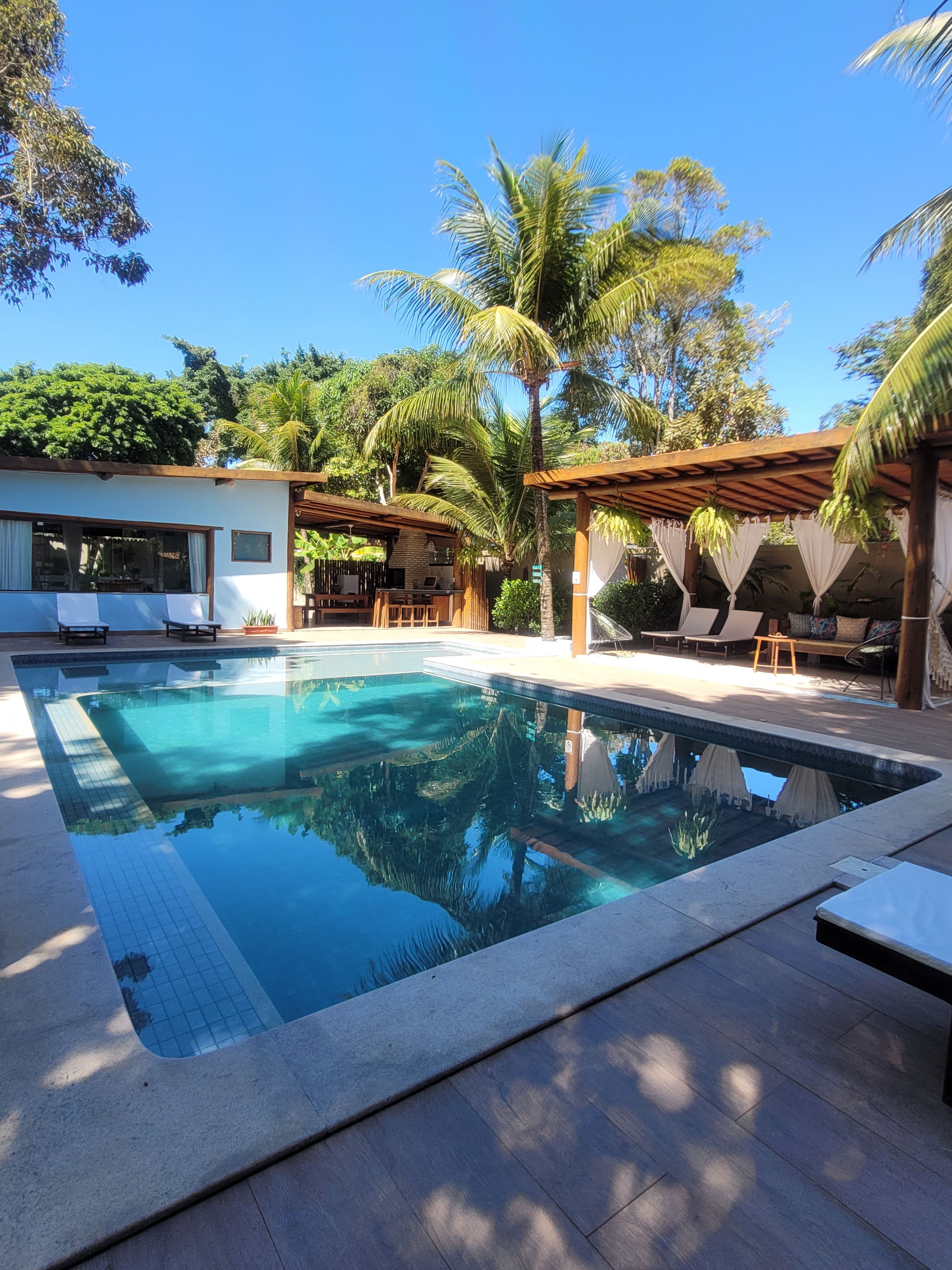 Piscina retangular com borda ladrilhada, rodeada por um deck de madeira e uma área coberta com mesas e cadeiras.