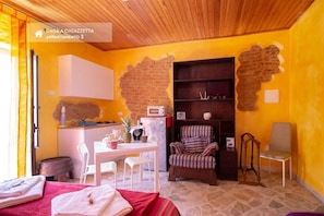 A cozy room with a brick wall, a wooden ceiling, a dining table with chairs, a bookshelf, and a small kitchen area. - House in Chiazzetta (Castelbuono)